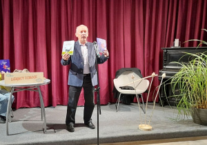 Pisarz stoi na scenie i prezentuje własne książki dla dzieci. Z lewej strony znajduje się stolik z tablicą "Latający Dziadek, a z prawej fotel, kwiat, fortepian.